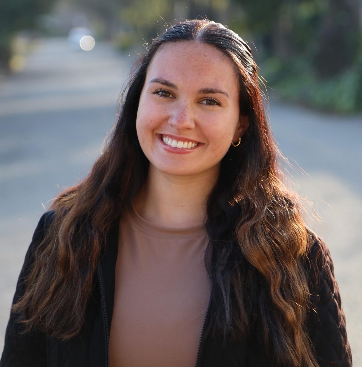 Colored portrait of a female with black hair with light brown highlights. She is smiling and staring at the camera. She is wearing a maude colored shirt with a black coat. A blurred background of a street is behind her. The photo was taken by her friend, Helen.