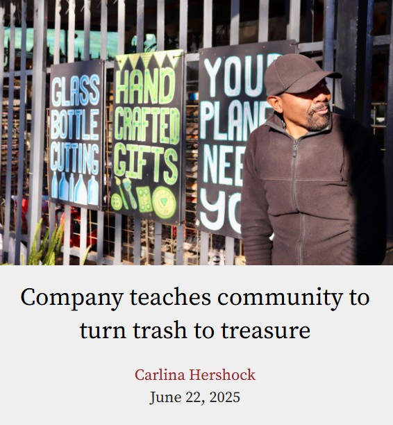 A photo of an African American man up against a face and looking to his left. He is standing outside of a shop called 'Upcycle.' The man is dressed in all black and has a black hat on. Behind him are signs with blue and green writing. The sun is shining down on him.